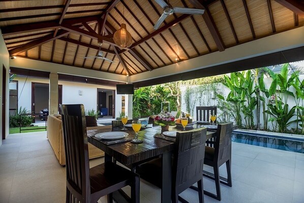 An outdoor dining area with a wooden table and chairs, surrounded by lush greenery and a swimming pool. - 5 Bdr -Last Minute Deal 50%+ OFF!!! (Legian)