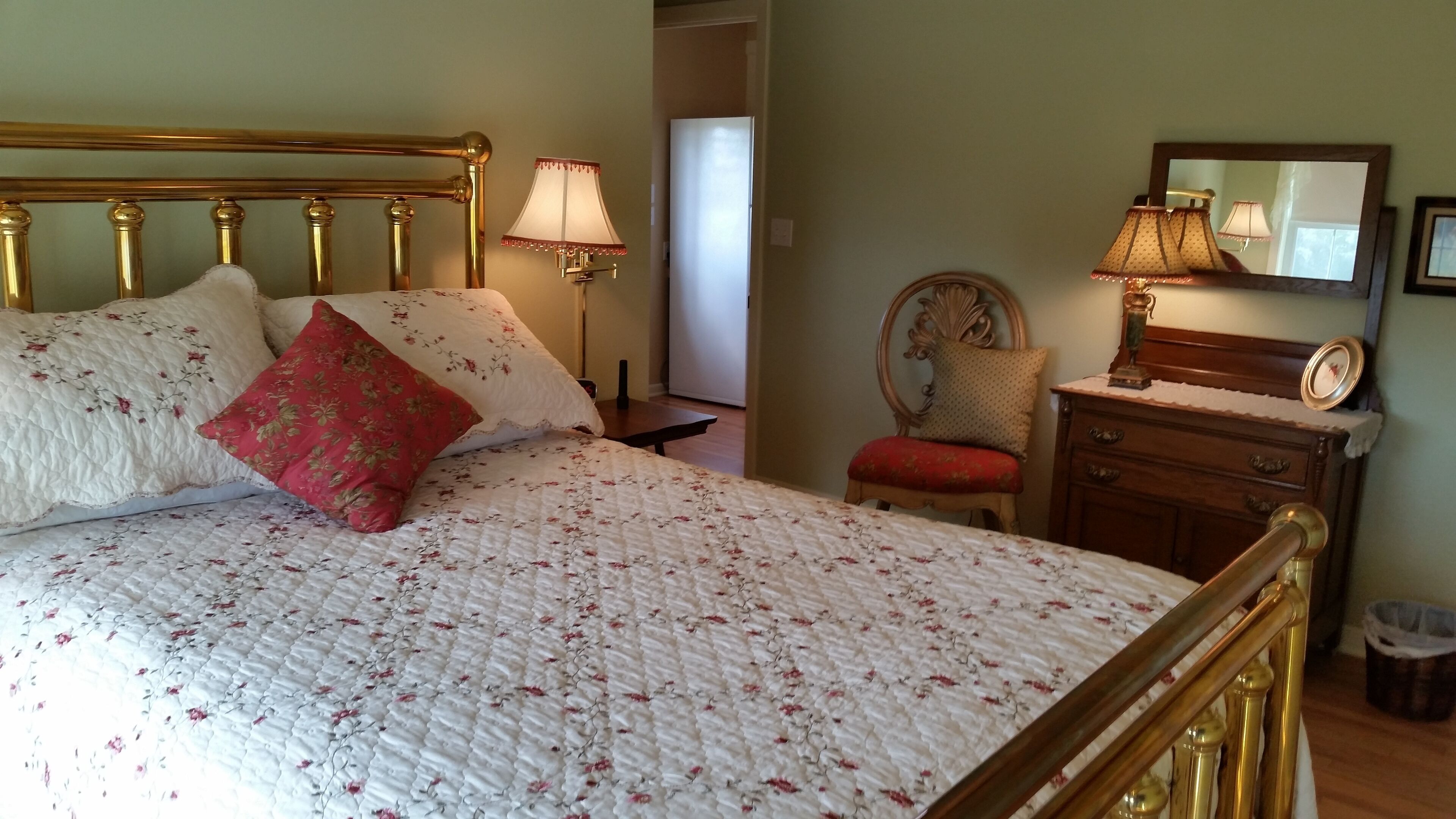 En pænt redt seng med et blomstret quilt, en rød pude og en guldramme om sengegærdet. Der er et trænatbord med en lampe, en stol med en rød pude og en trækommode med et spejl.