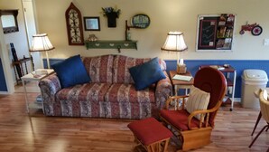 A living room with a patterned sofa, two blue cushions, a wooden rocking chair with a red cushion, a small table with a lamp, and a wall-mounted shelf with decorative items.