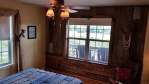 A room with wooden walls, a large window with a view of the outdoors, a ceiling fan, and a bed with a blue bedspread.