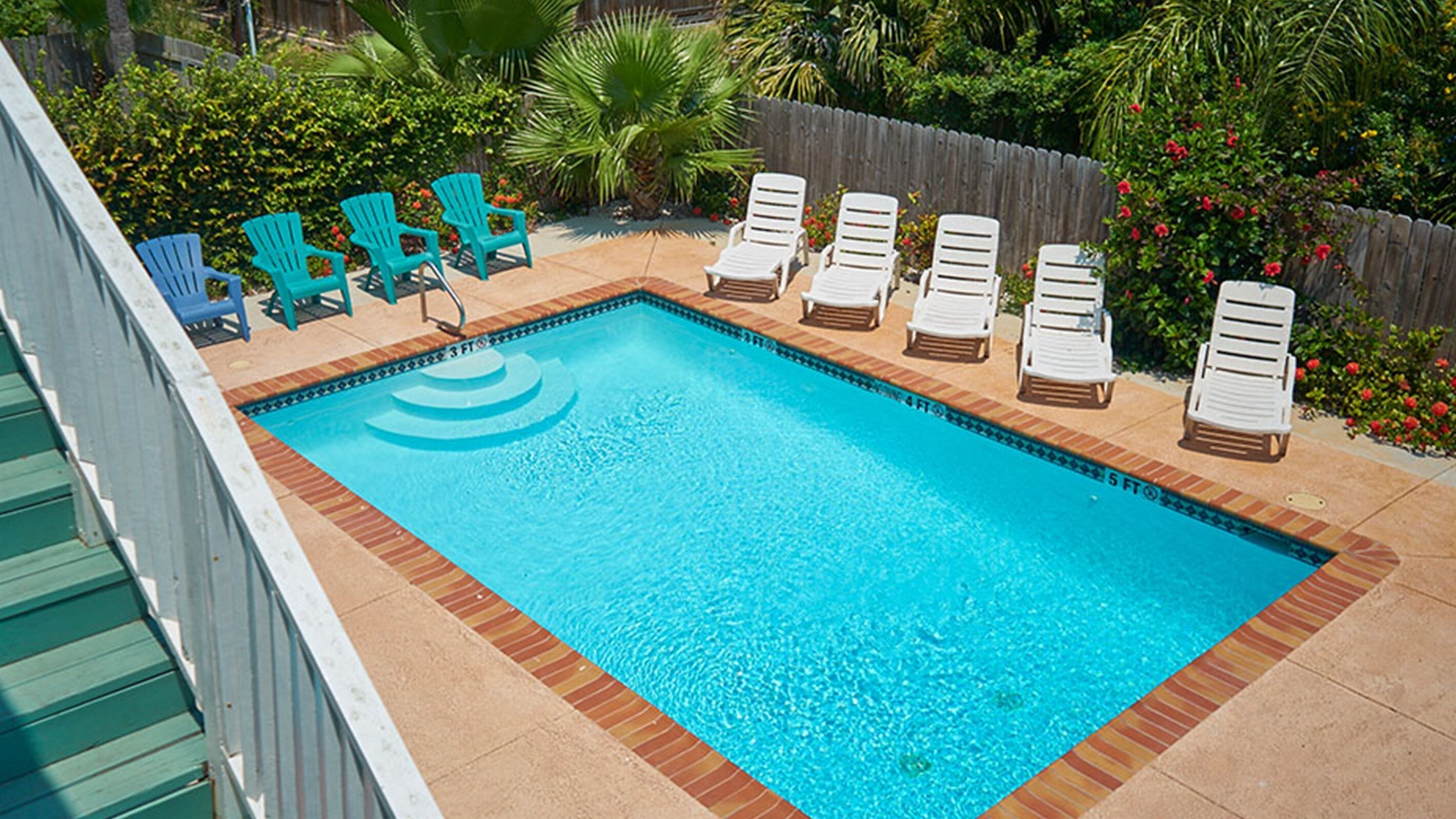 Una piscina rectangular con un deck de azulejos, rodeada de sillones de descanso blancos y sillas Adirondack azules.