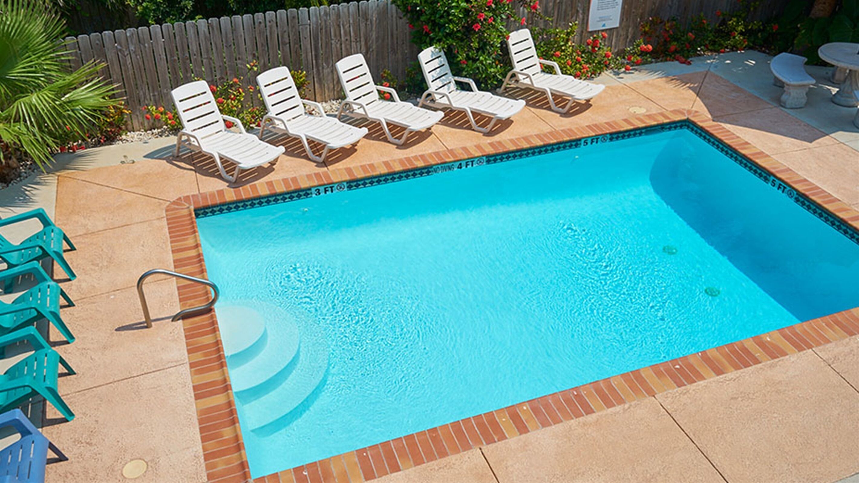 Una piscina rectangular con borde de ladrillo y solárium con baldosas. Hay sillas de descanso blancas y una mesita en el solárium.