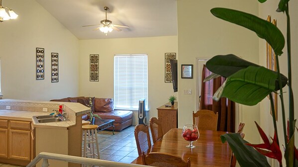 A modern kitchen with a dining area, a ceiling fan, and a large window with blinds.