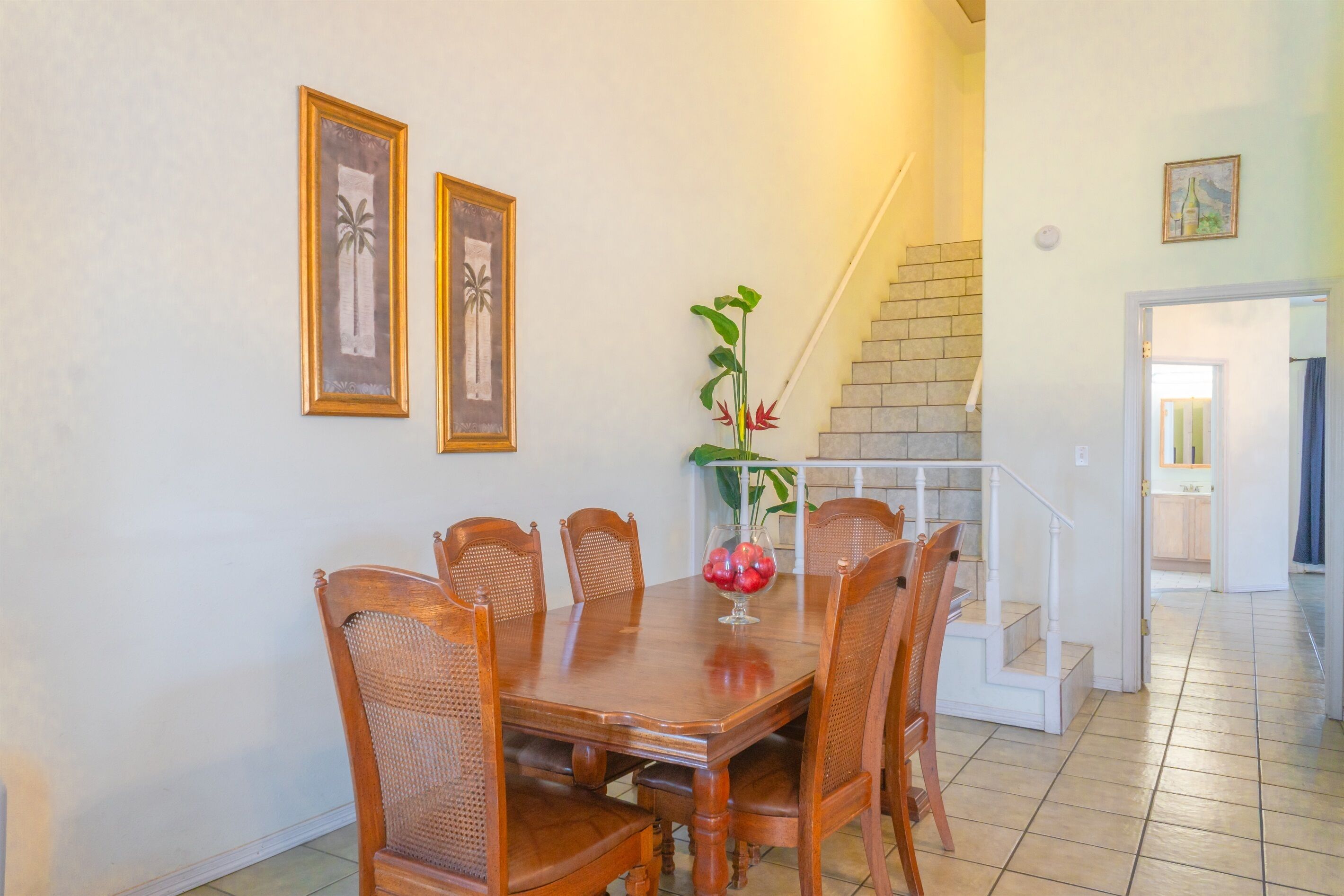 Uma sala de jantar com uma mesa e cadeiras de madeira, um vaso com flores vermelhas e duas molduras na parede.