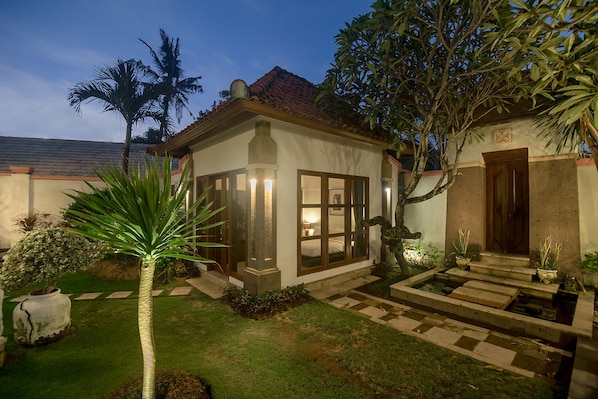 A small, well-lit house with a tiled roof, surrounded by a garden with a palm tree and a water feature.