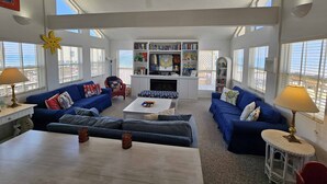 A spacious living room with a high ceiling, large windows, a ceiling fan, and a fireplace. - enJoy | gulfside, great price, open starting 8/8 (Dauphin Island)