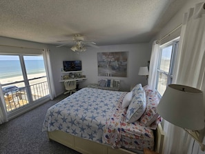 Quarto com cama, escrivaninha, cadeira, televisão e vista para a praia.