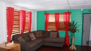 A living room with a sectional sofa, red curtains, a small wooden table, and a potted plant. - Cali Cottages (Negril)