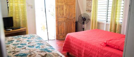 A bedroom with a bed, a wooden wardrobe, a desk with a television, and a fan.