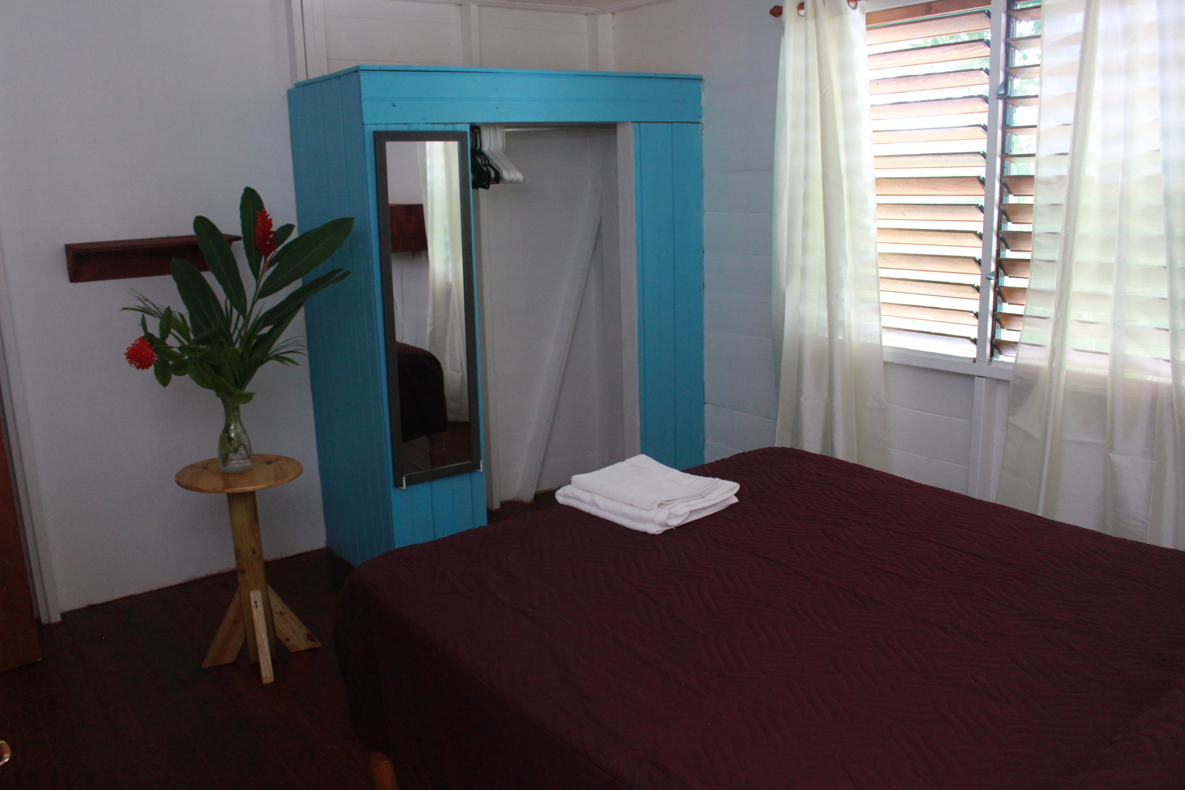 A room with a bed, a mirror, a small table with a plant, and a window with blinds.