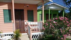 A two-story house with a porch, a small garden with pink flowers, and a concrete pathway leading to the entrance. - Cali Cottages (Negril)