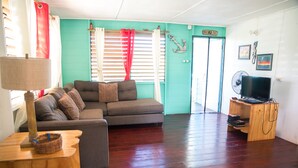 A living room with a sofa, a wooden side table, a lamp, a television on a wooden stand, and a door with a sign. - Cali Cottages (Negril)
