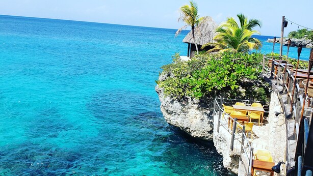 A coastal view with clear turquoise waters, a thatched-roof structure, and outdoor seating. - Cali Cottages (Negril)