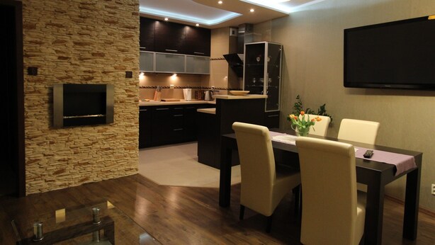 A modern kitchen with dark cabinets, a stone accent wall, and a flat-screen TV mounted above the counter. - Torun Apartment Modern apartment in the Torun city center (Jakubskie Przedmiescie)