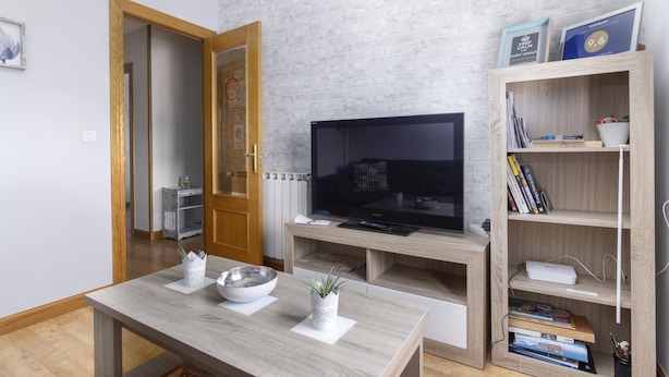A modern living room with a flat-screen TV, wooden shelves, a dining table, and a kitchen area visible through an open door.