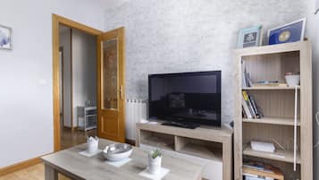 Una sala de estar moderna con un televisor de pantalla plana, estanterías de madera, una mesa de comedor y una cocina visible a través de una puerta abierta.