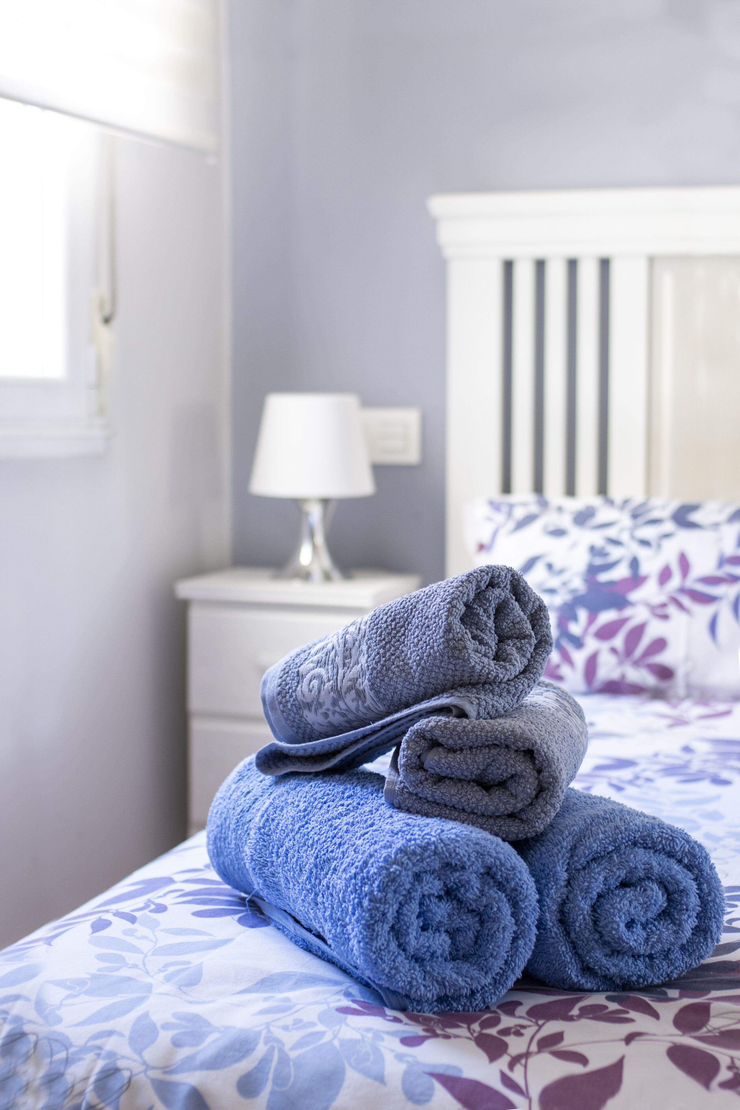 Una cama bien hecha con toallas enrolladas, una mesita de noche blanca con lámpara y un cojín estampado con motivos florales.
