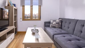 A modern living room with a grey sofa, a wooden coffee table, a TV on a wooden stand, and a window with blinds.