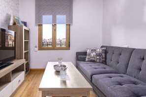Una sala de estar moderna con un sofá gris, una mesa de centro de madera, un televisor sobre un mueble de madera y una ventana con persianas.