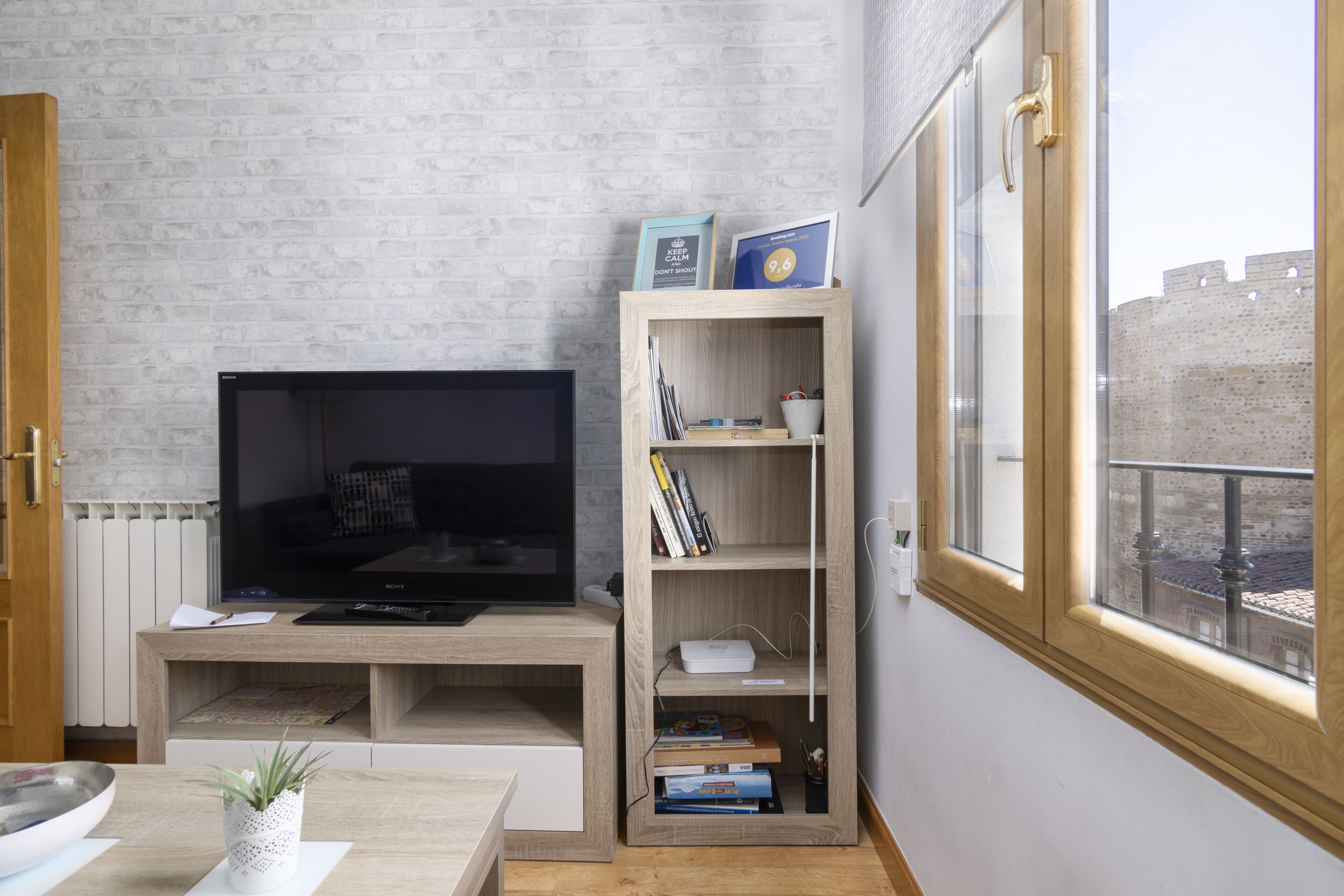 Una sala moderna con una televisión de pantalla plana, estanterías de madera con libros, una planta pequeña y vistas a un edificio histórico a través de una ventana.