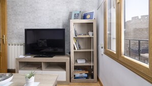 A modern living room with a flat-screen TV, wooden shelving with books, a small plant, and a view of a historic building through a window.