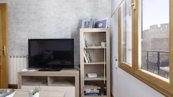 Una sala moderna con una televisión de pantalla plana, estanterías de madera con libros, una planta pequeña y vistas a un edificio histórico a través de una ventana.