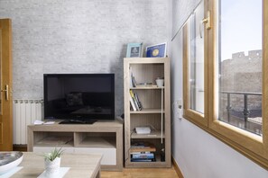 Una sala moderna con una televisión de pantalla plana, estanterías de madera con libros, una planta pequeña y vistas a un edificio histórico a través de una ventana.