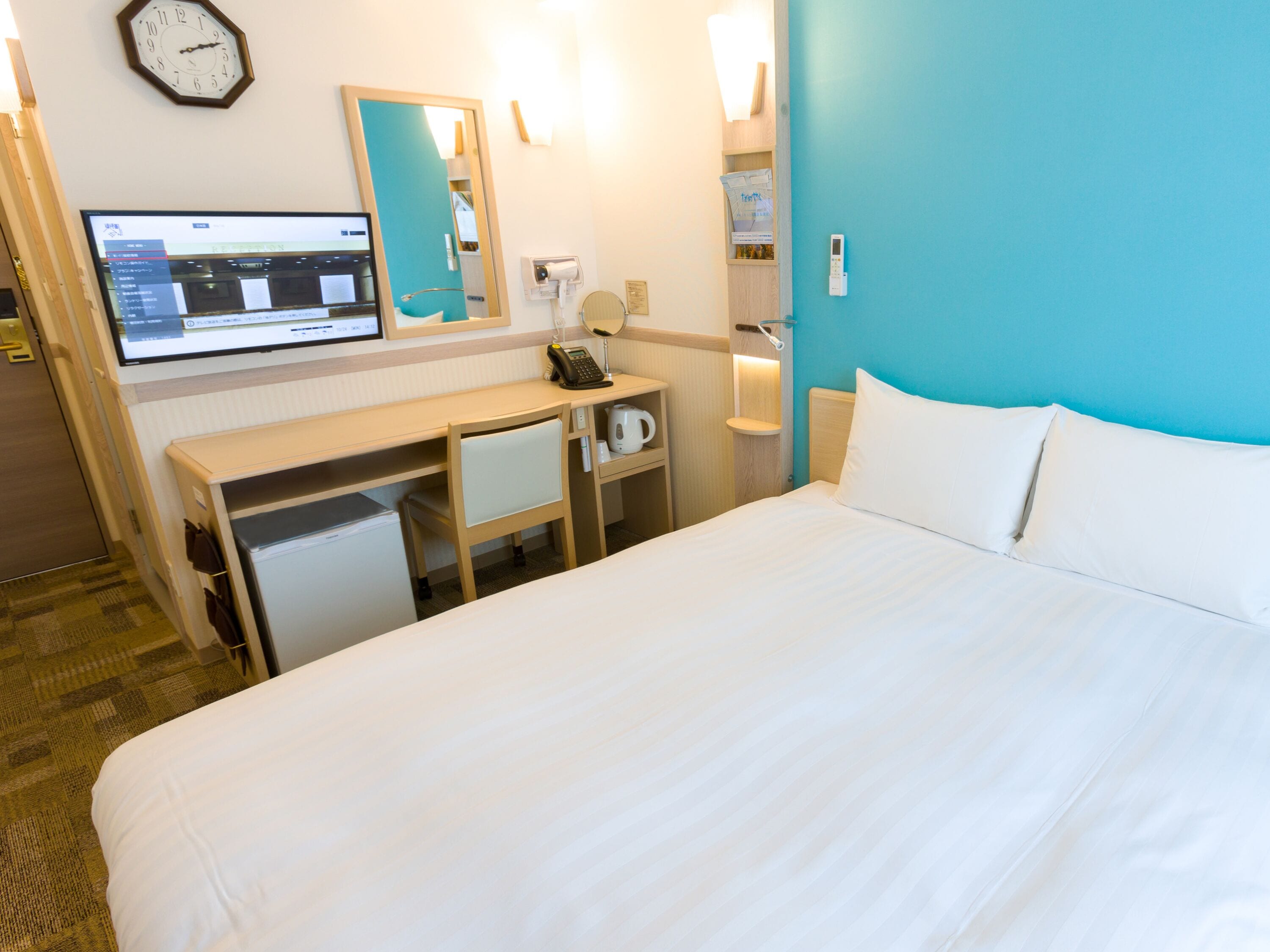 A hotel room with a bed, a desk, a computer, a mirror, and a clock on the wall.