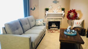 A living room with a grey sofa, a wooden coffee table, a fireplace with a 'Mermaids Welcome' sign, and a decorative wall with fish and mermaid figures. - Seaside Villa by the Oso Bay Comfortable Bed! (Corpus Christi)