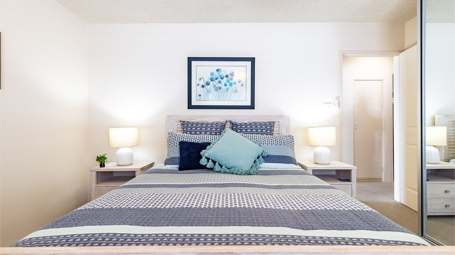 A neatly arranged bedroom with a large bed, two bedside tables with lamps, a framed artwork on the wall, and a door leading to another room.
