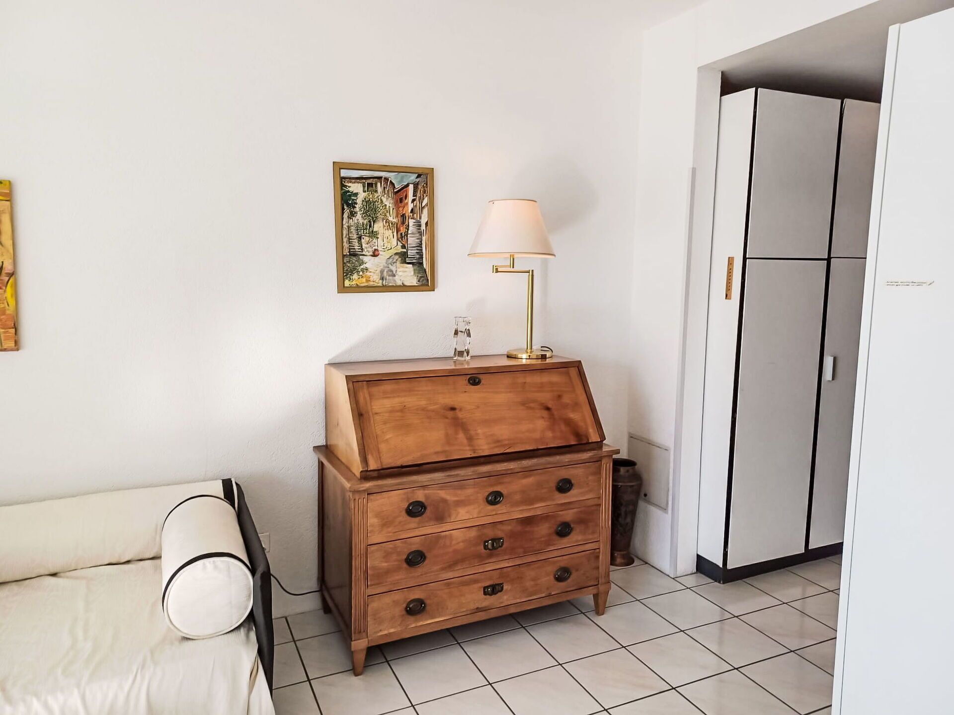 A room with a bed, a wooden chest of drawers, a lamp, a painting, and a white wardrobe.
