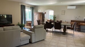 A spacious living room with a large sofa, a dining table with chairs, a television, and a piano. - Villa avec jardin et piscine (Fontvieille)