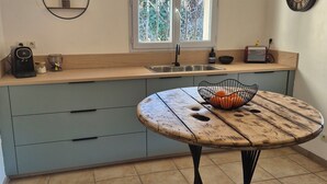A modern kitchen with a round wooden table, light blue cabinets, a window with a view, and pendant lighting. - Villa avec jardin et piscine (Fontvieille)
