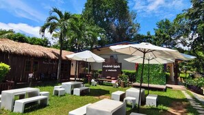 An outdoor seating area with concrete tables and umbrellas, surrounded by greenery and a thatched-roof building. - Ban Kiang Fah Resort Chongmek (Sirindhorn)