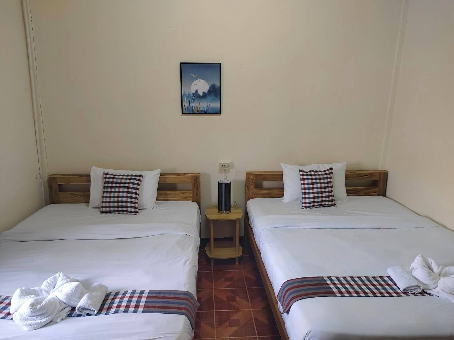 Une chambre d’hôtel avec deux lits, chacun agrémenté d’un coussin à carreaux et d’une couverture rayée. Il y a une table de chevet avec une lampe entre les deux lits, et un tableau représentant une lune et des nuages est accroché au mur.