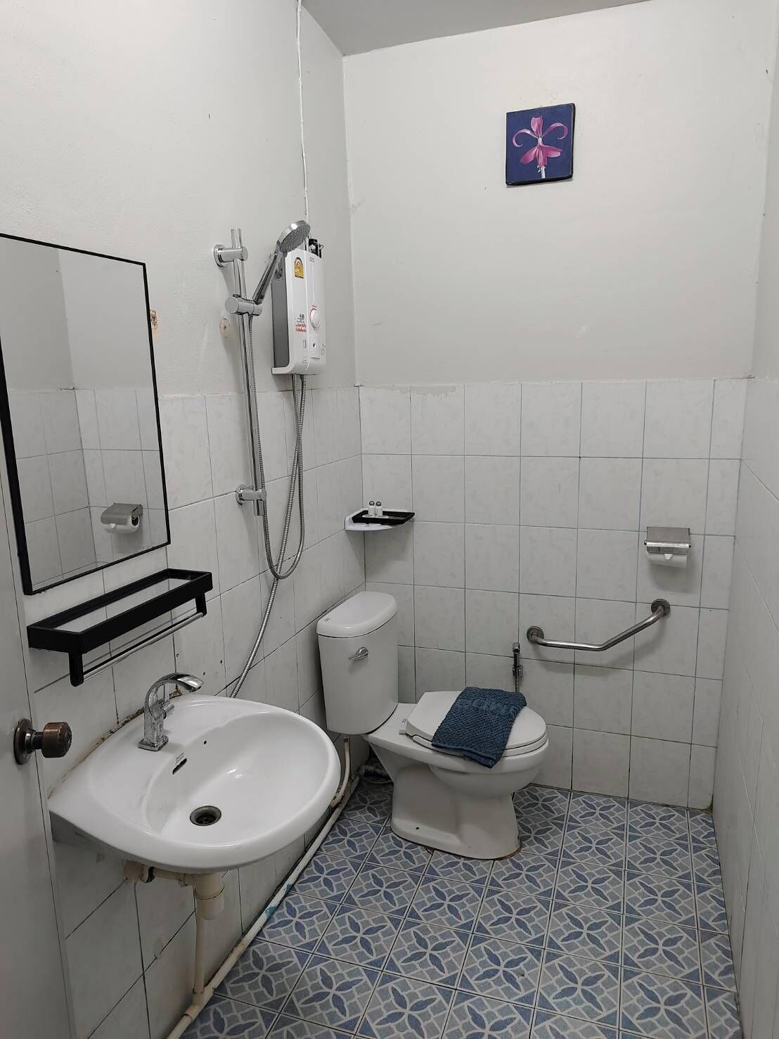 Un baño pequeño con un lavamanos, inodoro y ducha blancos. Hay una toalla azul sobre el inodoro, un pasamanos y un estante pequeño.