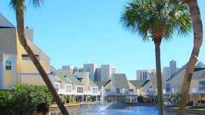 A waterfront hotel with a fountain, palm trees, and a view of the water. - Modern Sandpiper Retreat: 2 King Suites + Resort Golf, Pickleball & Dining (Destin)