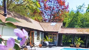 A swimming pool with a wooden deck, a covered outdoor seating area, and a house with a tiled roof. - Dog Friendly Cottage with Outdoor Swimming Pool, Tennis & Table Tennis (Chichester)