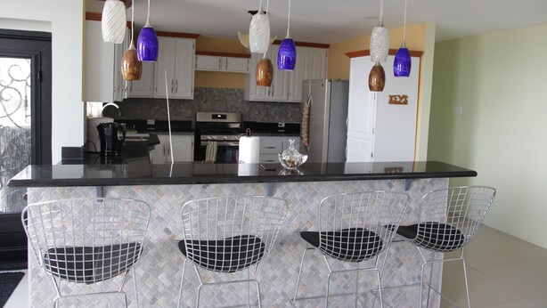 Una cocina con una isla central, lámparas colgantes y sillas de malla metálica.