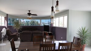 Una sala de estar moderna con una mesa de comedor, sillas, un ventilador de techo y grandes ventanas que ofrecen vistas al exterior.