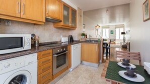 A modern kitchen with wooden cabinets, a built-in oven, a dishwasher, and a microwave. There is a dining area with a table and chairs.