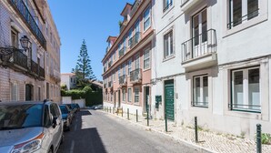 A narrow street lined with multi-story buildings, parked cars, and a cobblestone sidewalk.
