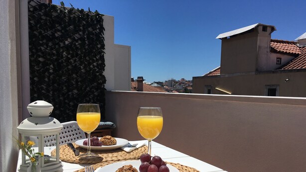 A table set with a plate of cookies and grapes, two glasses of orange juice, a small vase with flowers, and a lantern on a balcony overlooking a cityscape.