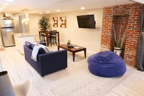 Una sala de estar moderna con un sofá, una mesa de centro y un sillón informal. Se ve una cocina con refrigerador y comedor al fondo.