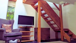 A living room with a TV, a sofa, wooden stairs, and a window with greenery outside. - Rural Mezzanine Studio with Shared Swimming Pool, Tennis, Table Tennis & zip wire (Chichester)