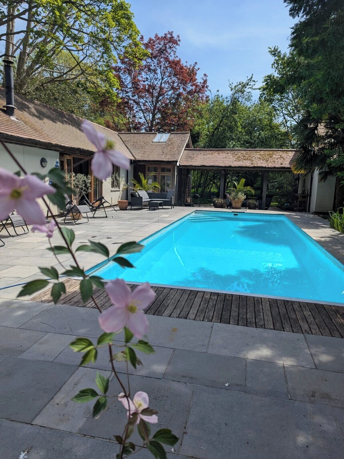 A rectangular swimming pool with a wooden deck surrounding it, a covered seating area with chairs, and a building with a tiled roof in the background.