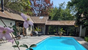 A rectangular swimming pool with a wooden deck surrounding it, a covered seating area with chairs, and a building with a tiled roof in the background. - Rural Mezzanine Studio with Shared Swimming Pool, Tennis, Table Tennis & zip wire (Chichester)