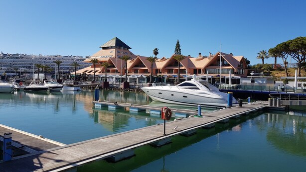 A marina with yachts docked, a wooden pier, and a building with a distinctive pyramid roof. - Walk to beach/marina, pool view, aircon, wifi, parking. Ideal Families/Groups. (Vilamoura)