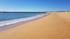 A wide sandy beach with gentle waves, a clear blue sky, and a rocky breakwater in the distance. - Walk to beach/marina, pool view, aircon, wifi, parking. Ideal Families/Groups. (Vilamoura)