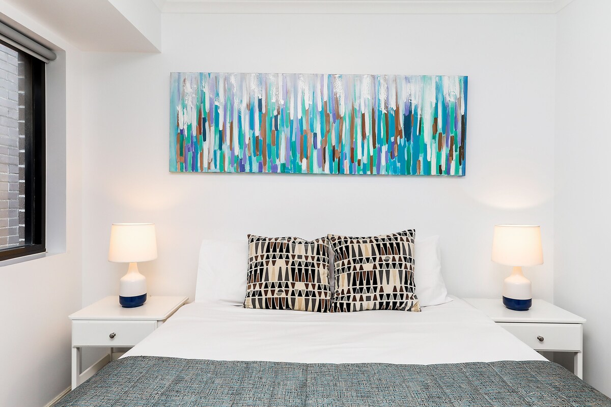 A bedroom with a large abstract painting, two bedside lamps, and patterned pillows.